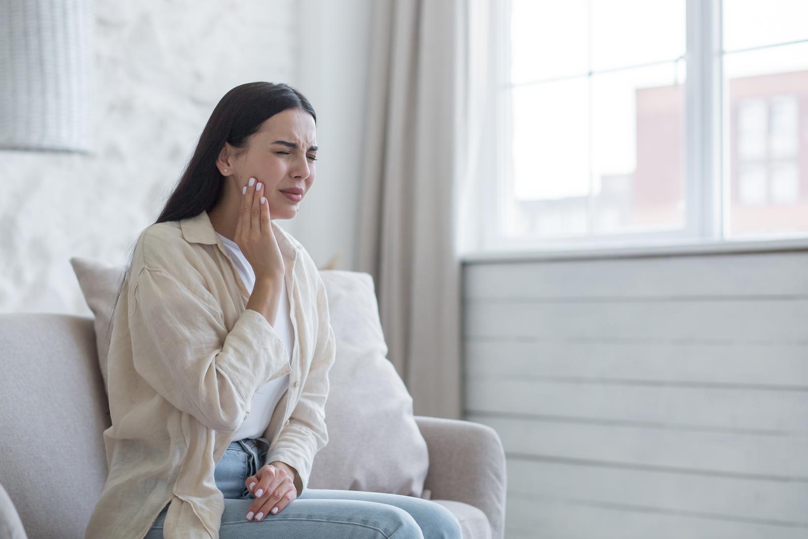 A woman is sick at home alone, has a severe toothache, a brunette is sitting on the sofa by the window, upset